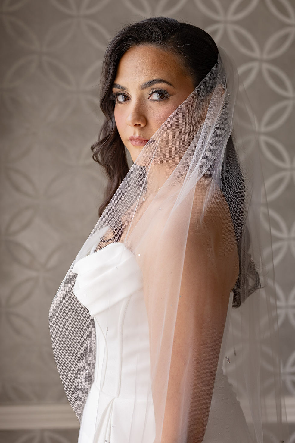 Bride in white dress with veil, gazing softly. Background has a geometric pattern. Mood is serene and elegant.