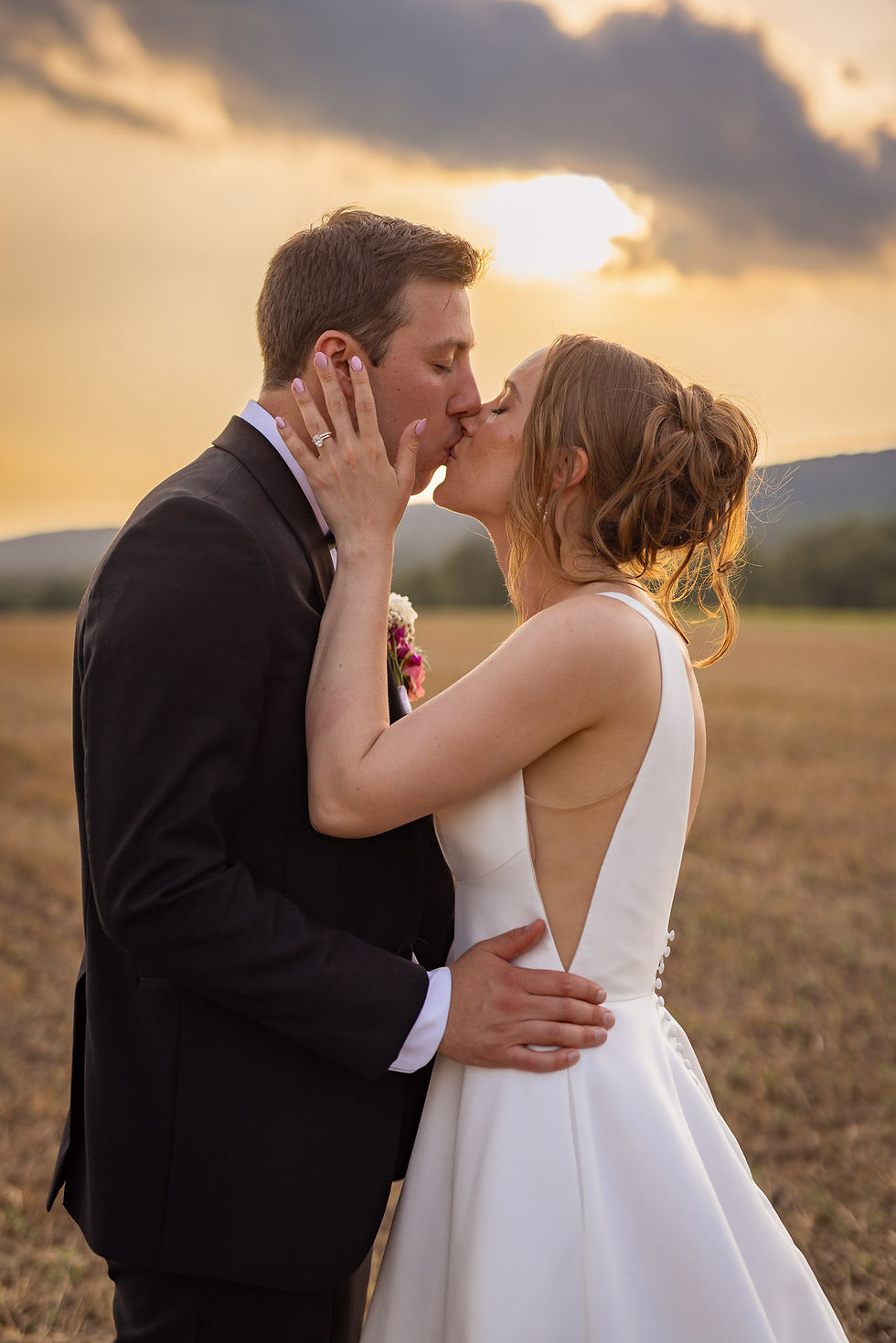 A couple kisses at sunset in an open field. The woman wears a white dress, the man a black suit, creating a romantic and warm mood.