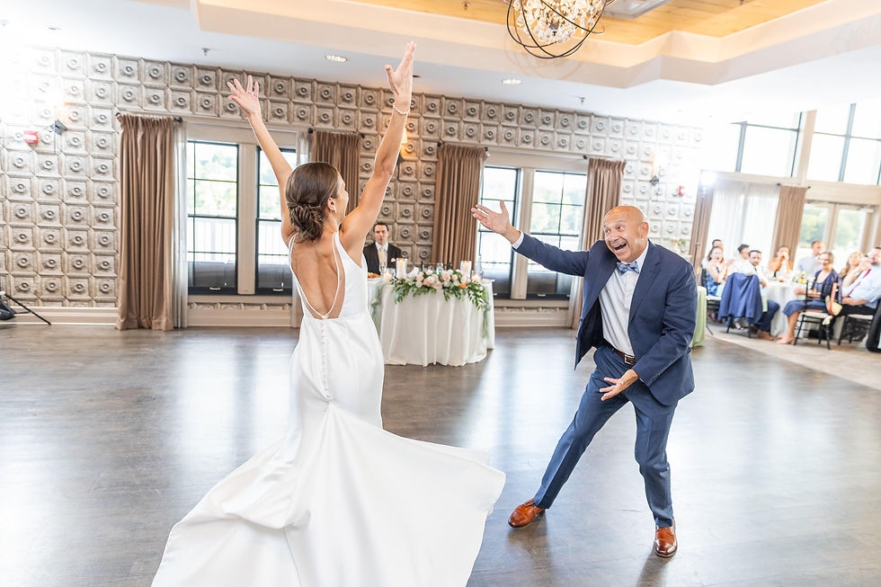 Bride in white gown dances with a smiling man in a blue suit at a bright wedding reception ballroom, guests watching nearby