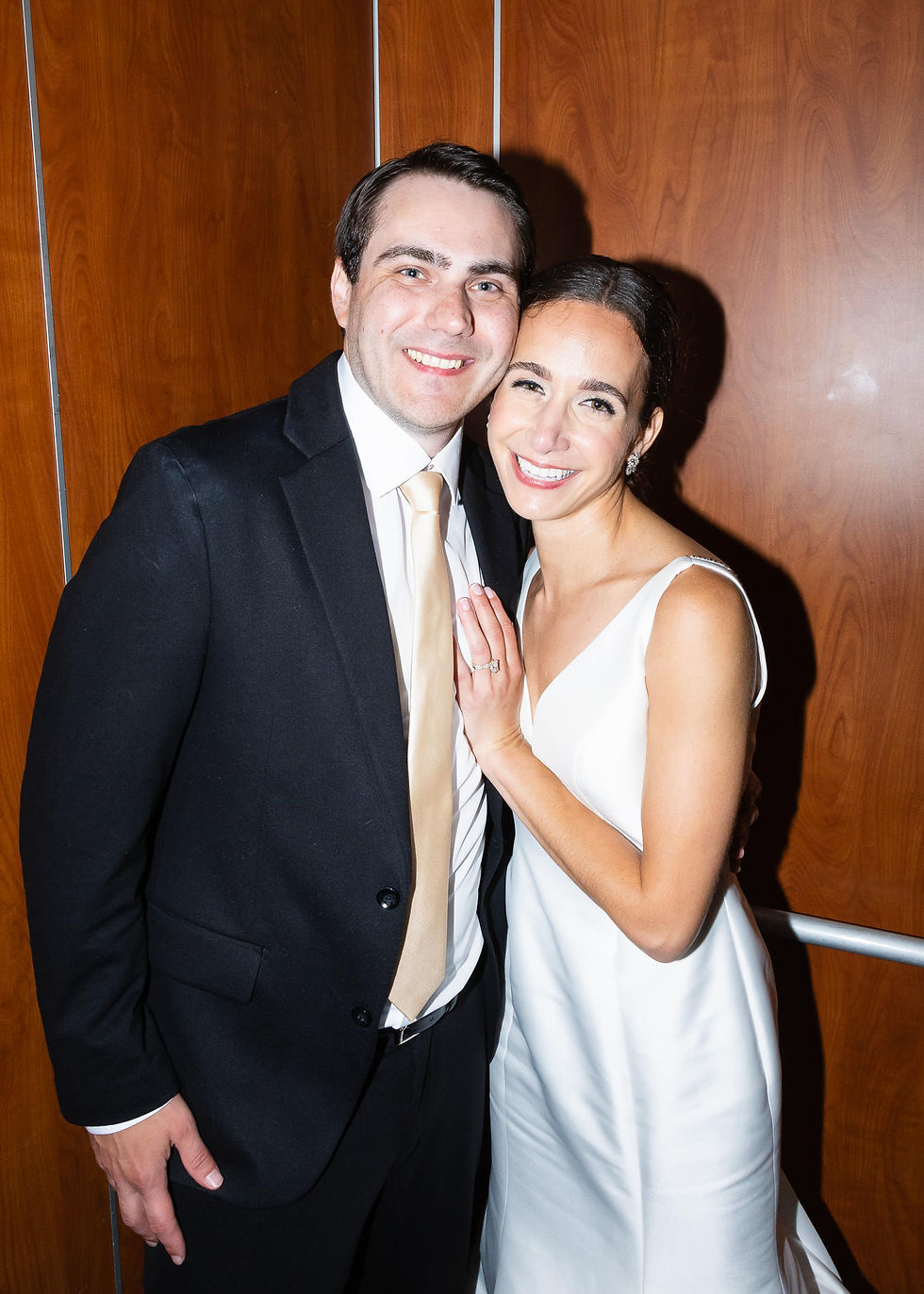 Smiling bride and groom in formal attire pose close together by a wood-paneled wall, her ring visible, warm and joyful mood