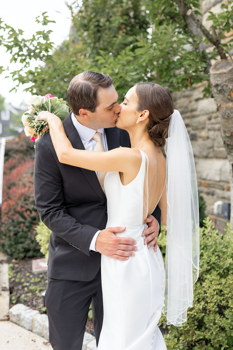 Bride in a white dress and veil kisses groom in a black suit outdoors, holding a bouquet amid greenery.
