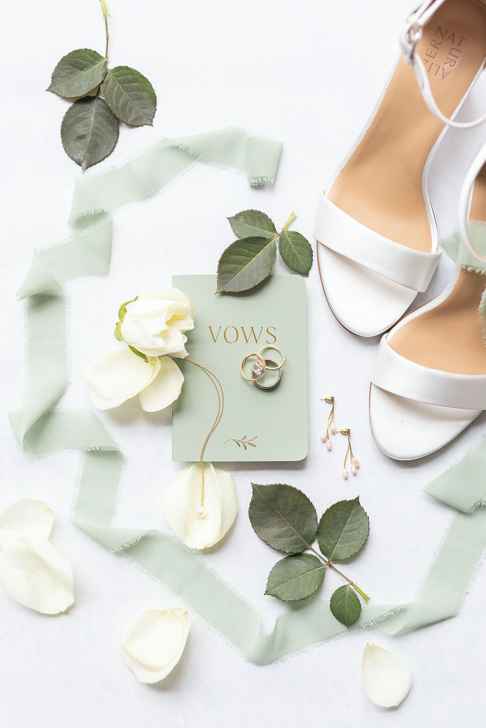 Wedding flatlay: gold rings on a green "Vows" booklet, pearl earrings, white shoes, and roses with leaves on a pale background.