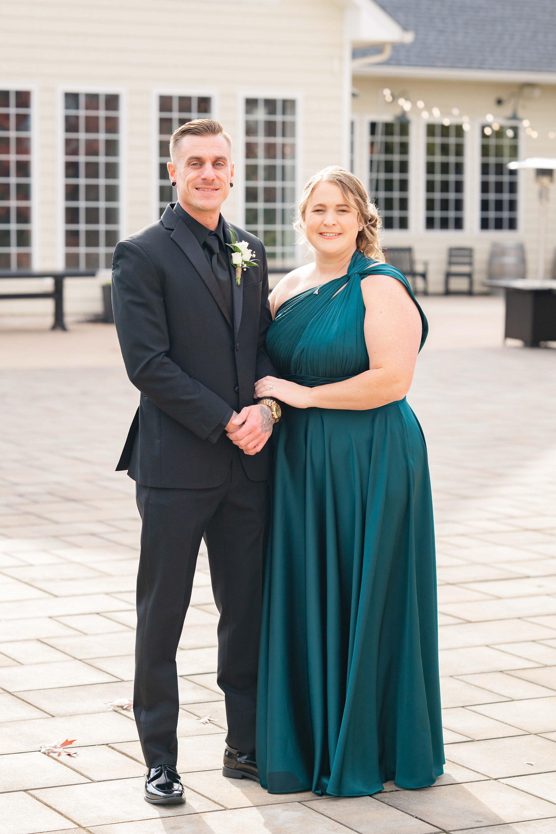 Man in black suit and woman in green dress stand on a patio in front of a cream building