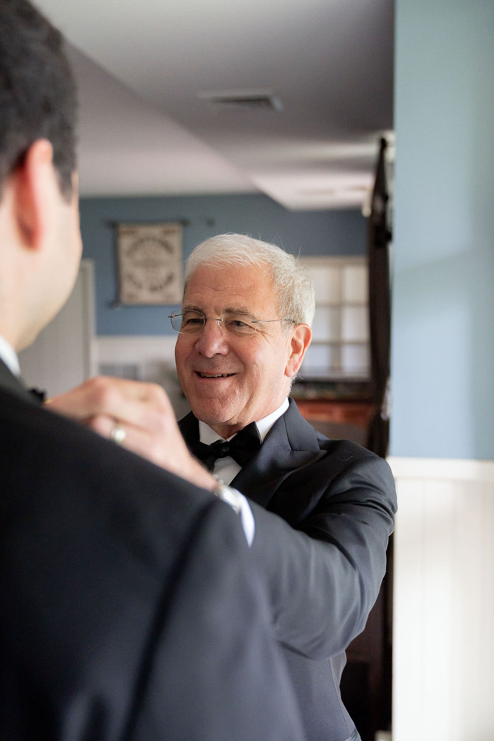 A father in glasses and a tuxedo with a black bow tie is seen over the shoulder of his son. He adjusts his son's bow tie as he smiles.