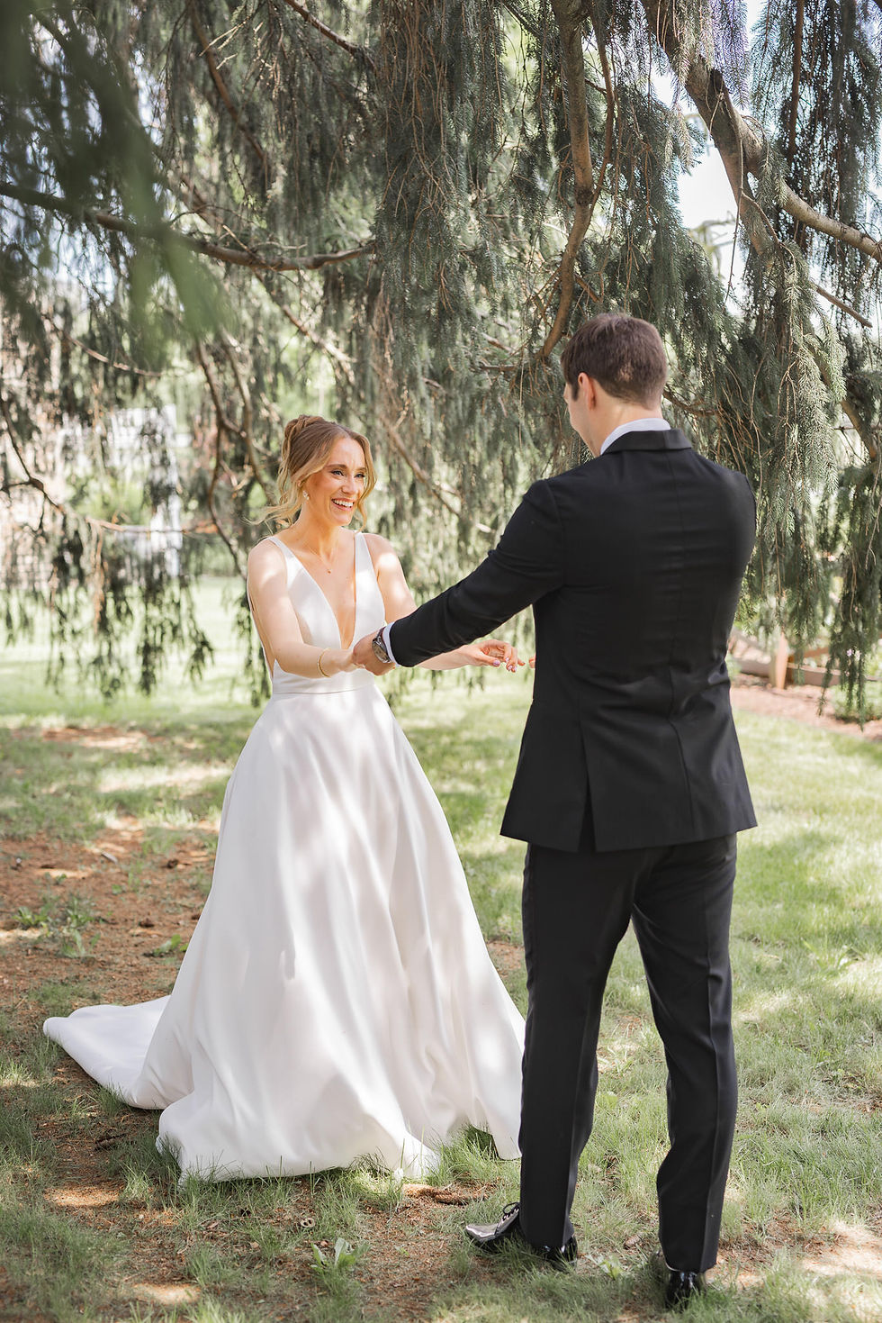 Bride in a white gown and groom in a black suit smile and hold hands under lush green trees in a serene outdoor setting.