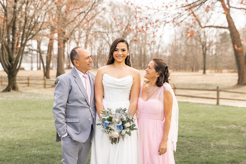 A bride in white with a bouquet stands between two smiling people. The setting is a grassy, wooded area with bare trees.