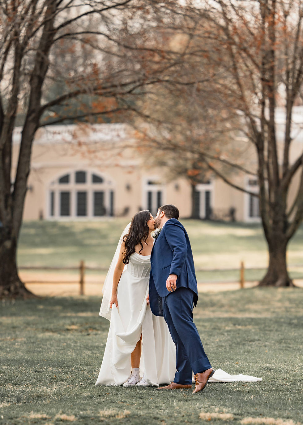 Bride in white dress and sneakers kisses groom in blue suit on lawn, in front of large house. Trees in background, autumn setting.