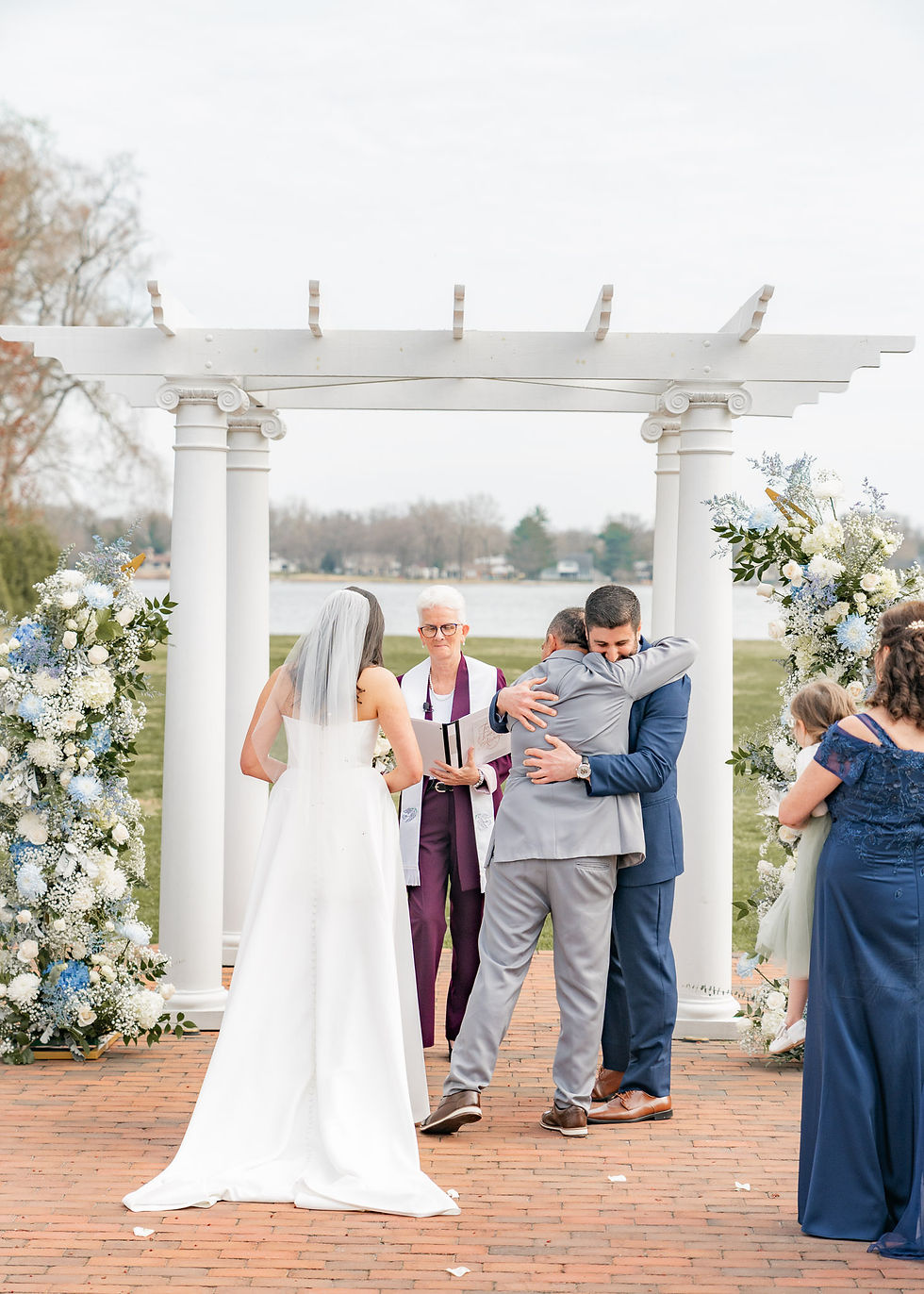 Bride in white dress, groom in navy suit hug another man under white pergola. Floral arrangements, officiant present, outdoor setting.