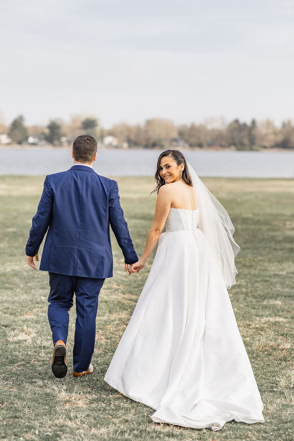 Bride in white gown and groom in blue suit hold hands, walking on grass by a lake. Bride glances back smiling, creating a joyful mood.