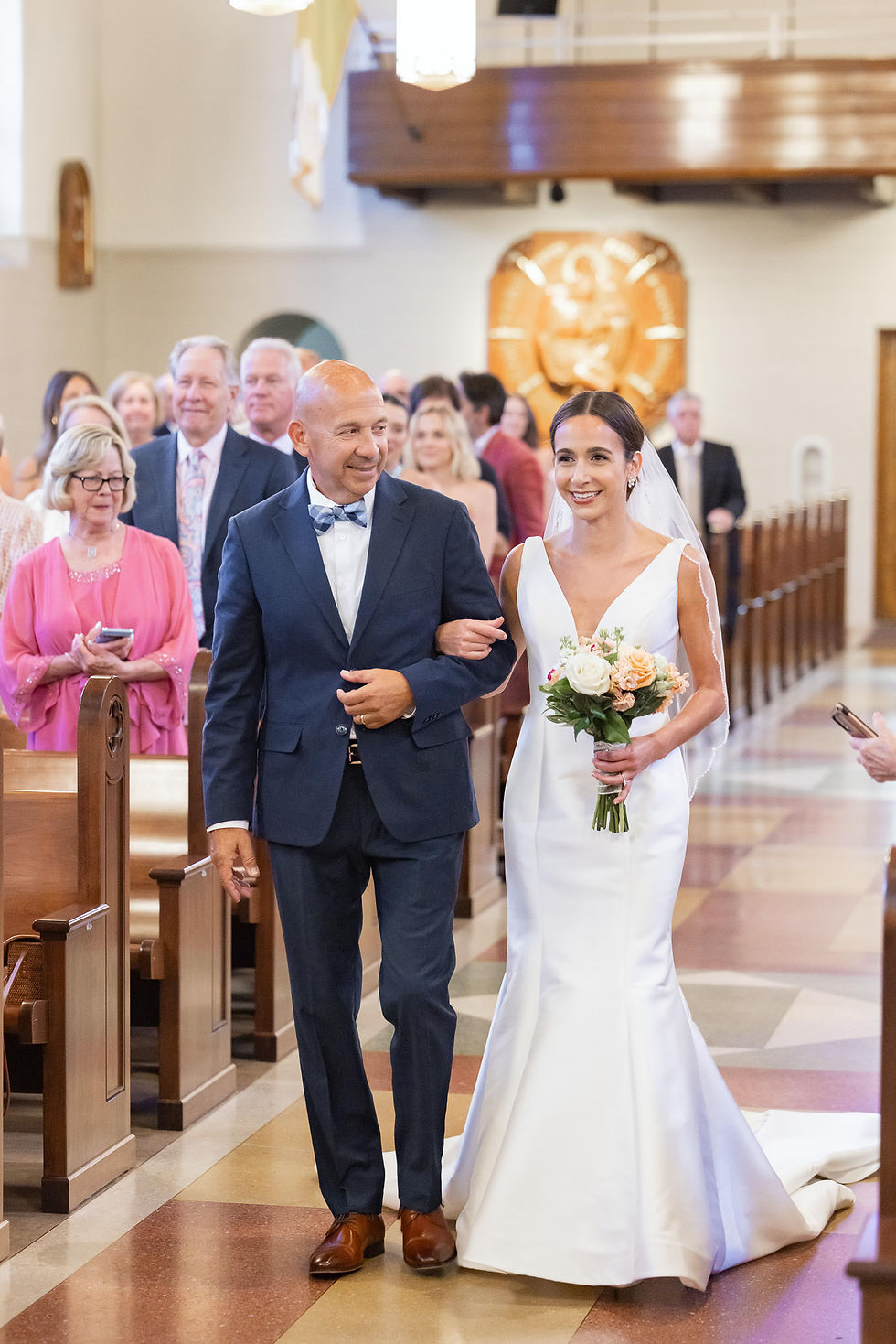 Bride in white gown walks arm-in-arm with a man down a church aisle, smiling, bouquet in hand, with guests watching.