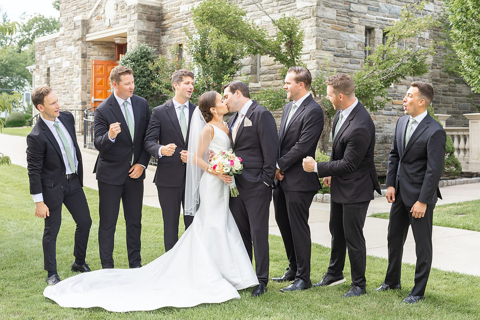 Bride and groom kiss on a lawn outside a stone building, surrounded by six groomsmen in black suits reacting with smiles.