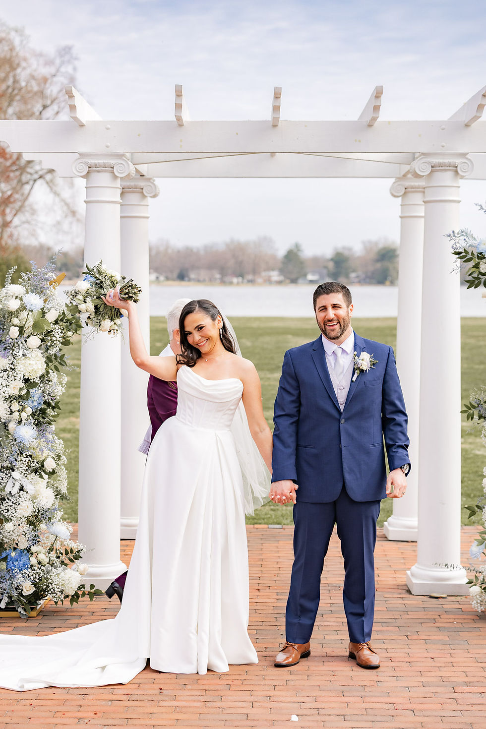 Bride and groom celebrate, holding hands under a white arbor decorated with flowers. Bride raises bouquet, both smiling, outdoors by a lake.