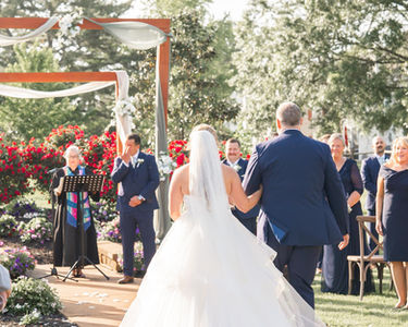 A groom in a blue suit wipes tears facing a bride in a white dress being walked down the aisle by an older man in a blue suit.  Guests smiling looking on in a jovial outdoor setting.