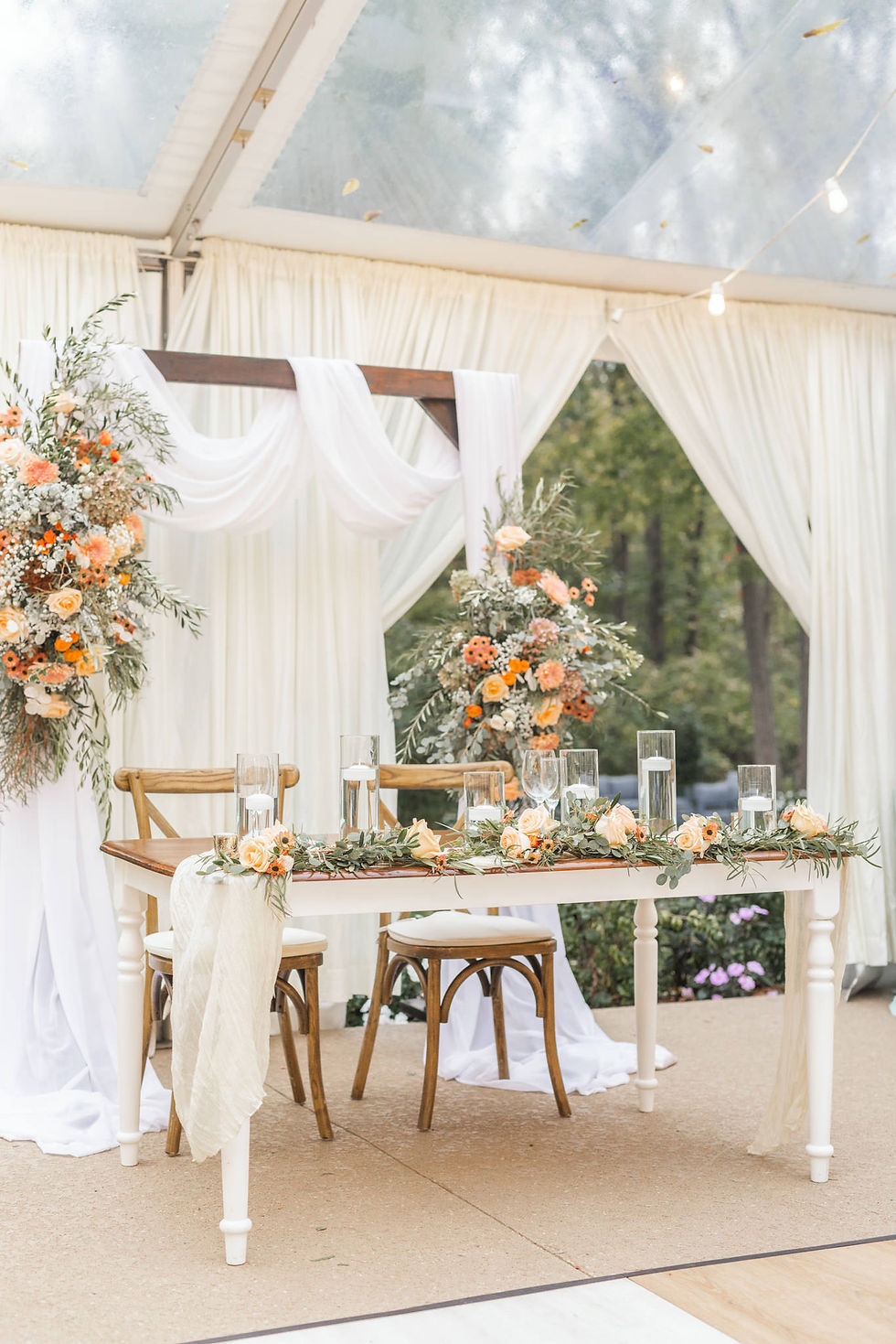 Elegant wedding table setup with floral arrangements in orange and white. Wooden chairs and draped white fabric in a tented outdoor setting.