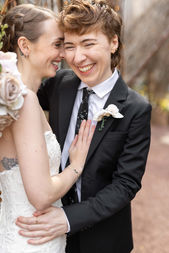 Woman in a black suit with an orchid boutonnière smiles broadly and holds another woman in a white dress who laughs and rests her hand on her chest.
