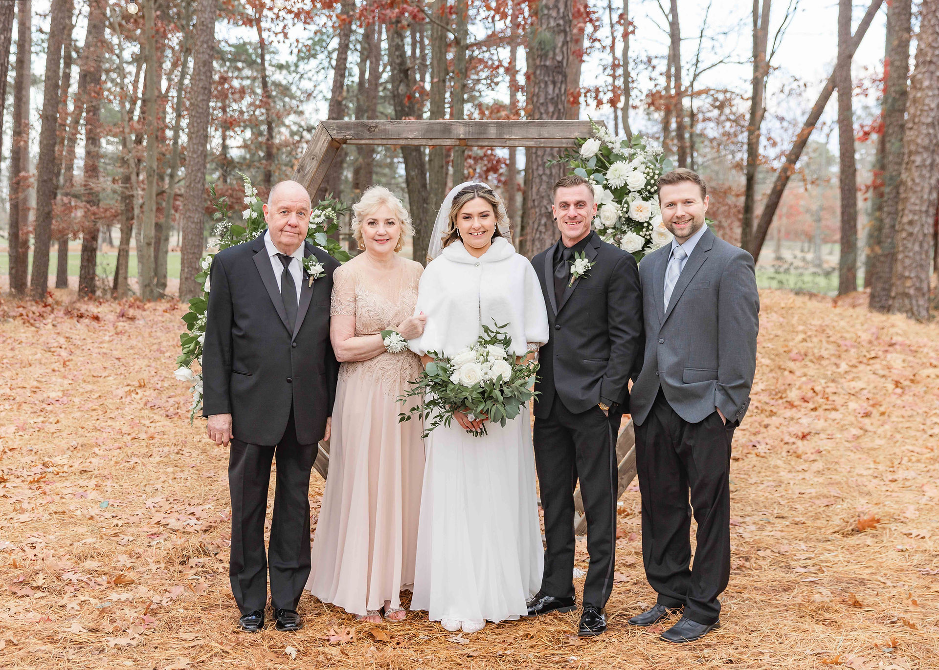Formally dressed parents and brother stand with bride and groom in front of floral arch in an autumn grove