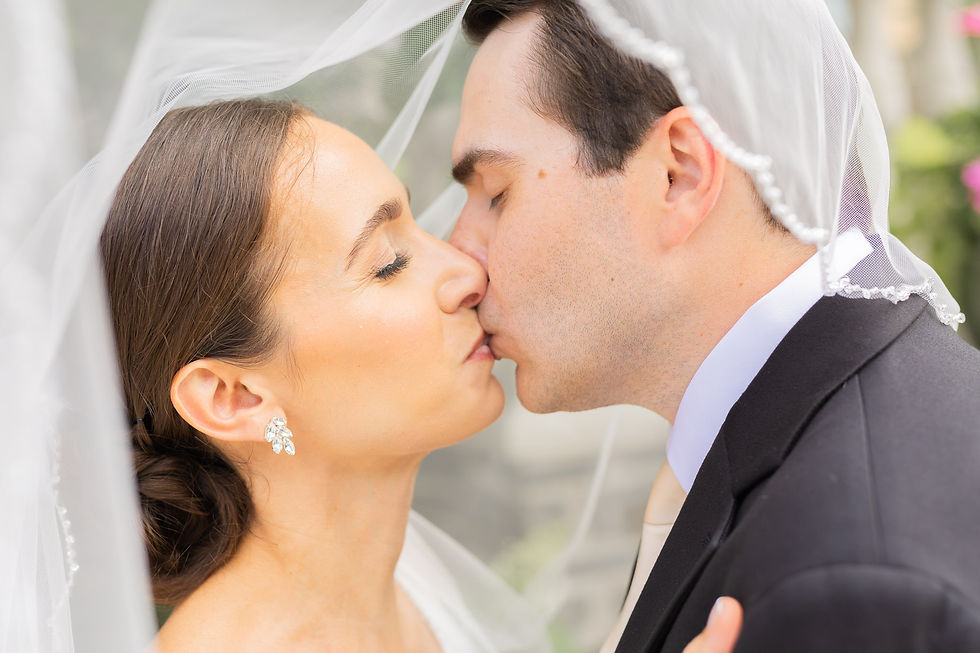 Bride and groom kiss under a sheer wedding veil, eyes closed, in a soft outdoor close-up with a romantic mood.