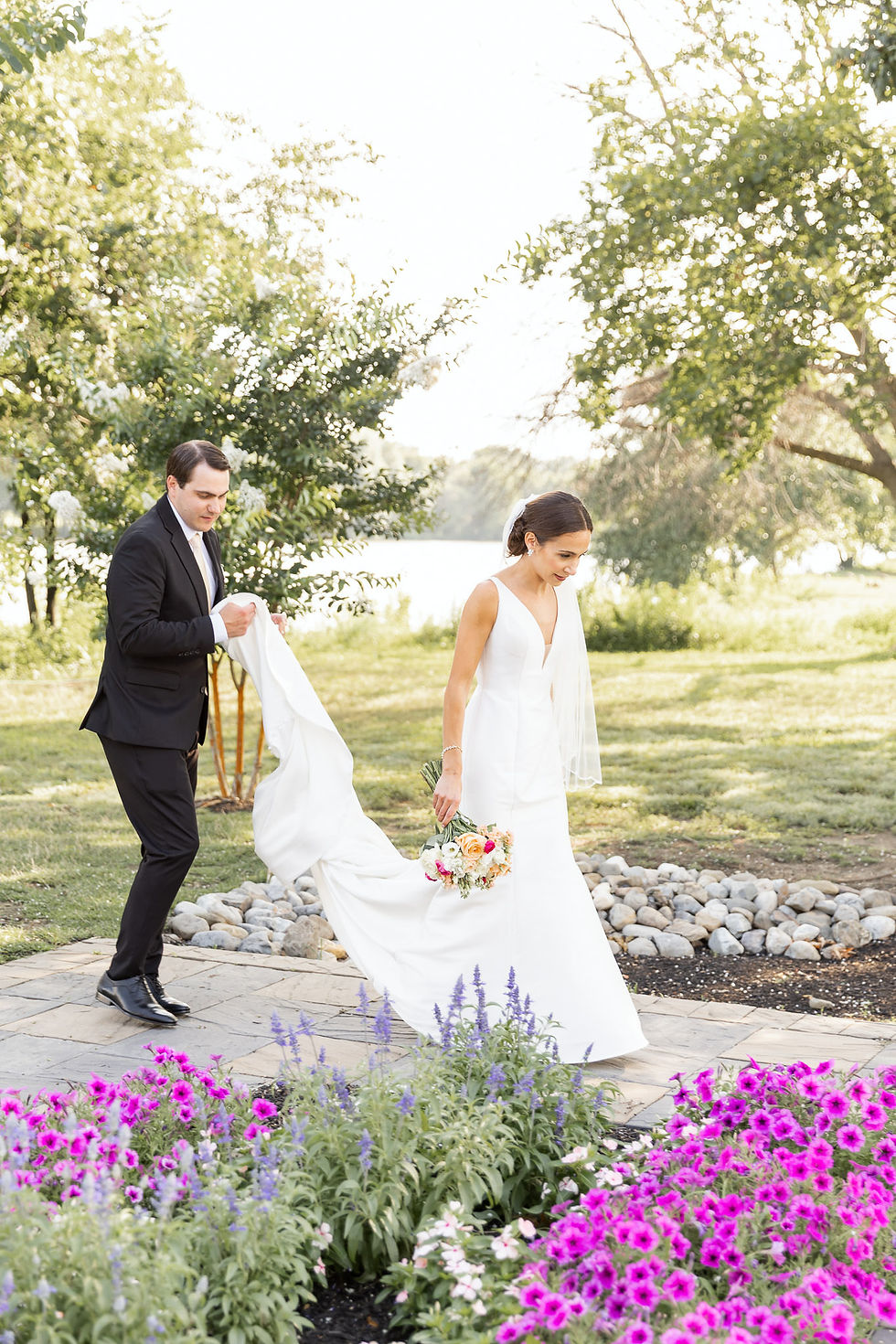 Bride in white gown walks through garden holding bouquet as a man in a black suit carries her train, with bright flowers around them