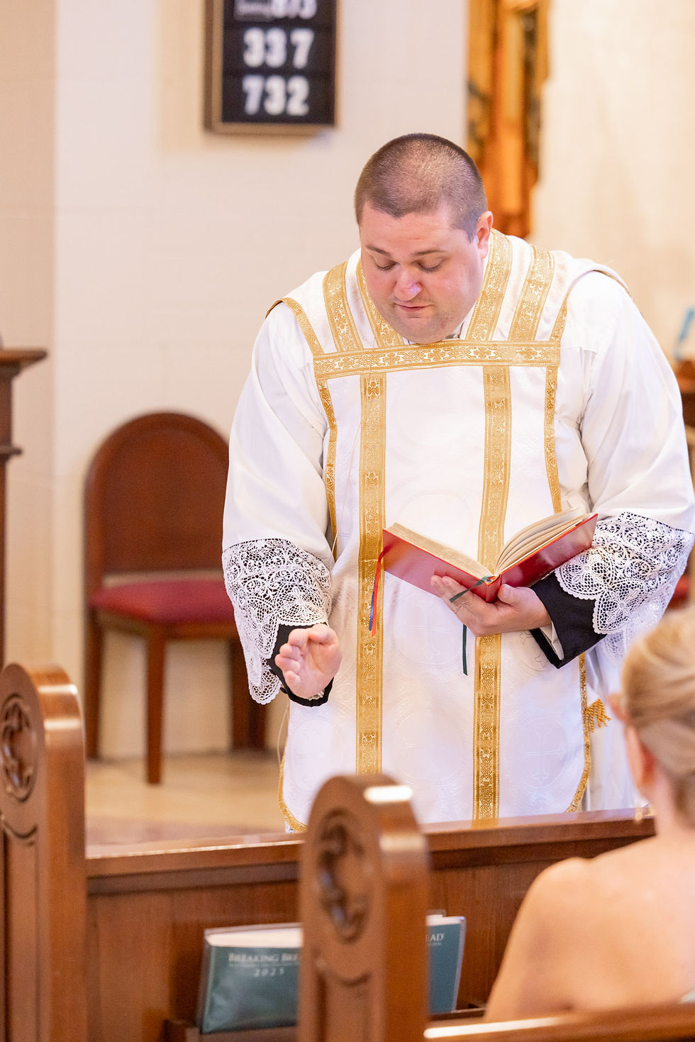 Priest in white-and-gold vestments reads from a red book in a church; hymn numbers 337 and 732 hang behind.