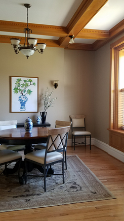 This dining room in a historic home has tall, beamed ceilings, neutral walls, and lots of natural light. A round black dining table is surrounded by cream-colored leather chairs with iron crossbacks.