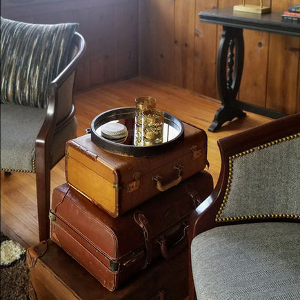 A rustic cabin interior has knotty pine paneling. Vintage leather suitcases serve as a side table between two nailhead chairs.
