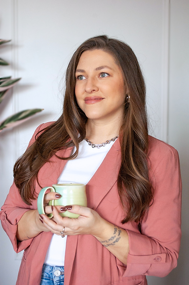 Alexa, smiling and looking off-camera, stands in front of a white wall wearing a pink linen blazer, a white t-shirt, and blue jeans, and holds a green and teal handmade mug from Alaska.