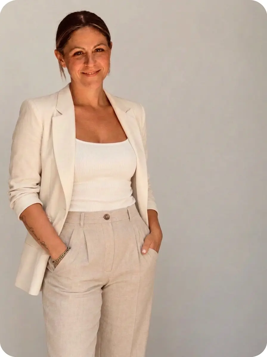Studio portrait photography of a woman dressed in a cream blazer and linen pants, hands in pockets, smiling confidently.