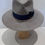Thumbnail: A face on view of an ash grey fedora with a black and multicolour ribbon around the base. A flat no curl brim and signature Marcel Rodrigues safety pin embellishment, showing the pinched crown. 