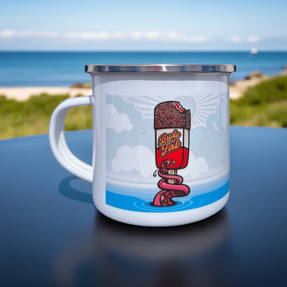 White enamel Bude Is Fab mug with octopus tentacle holding a lolly, displayed on a picnic table with the sea behind.