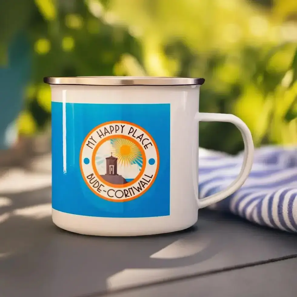 White enamel Bude Is My Happy Place mug outdoors on a picnic table with a blue and white striped tea towel