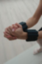 Close-up of clasped hands wearing black wrist weights during a Pilates session. The soft, neutral-toned background and light flooring provide a calming setting, while the person’s hands and wrists, adorned with neatly manicured nails, are the focal point. 這張特寫展示一雙戴著黑色手腕負重裝備的手,背景是柔和淺色調地板,增添了平靜氛圍。雙手交握,指甲修整整齊,成為畫面焦點。