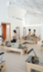 A pilates class in session at a bright and spacious studio. The instructor is guiding a group of women seated on reformer machines, demonstrating proper technique. The studio features high ceilings, modern equipment, and a clean, minimalist design.在明亮寬敞的工作室裡進行的普拉提課程。教練正在指導一組坐在普拉提滑輪機上的女性,演示正確的技術。工作室擁有高高的天花板、現代化的設備和乾淨的極簡設計。