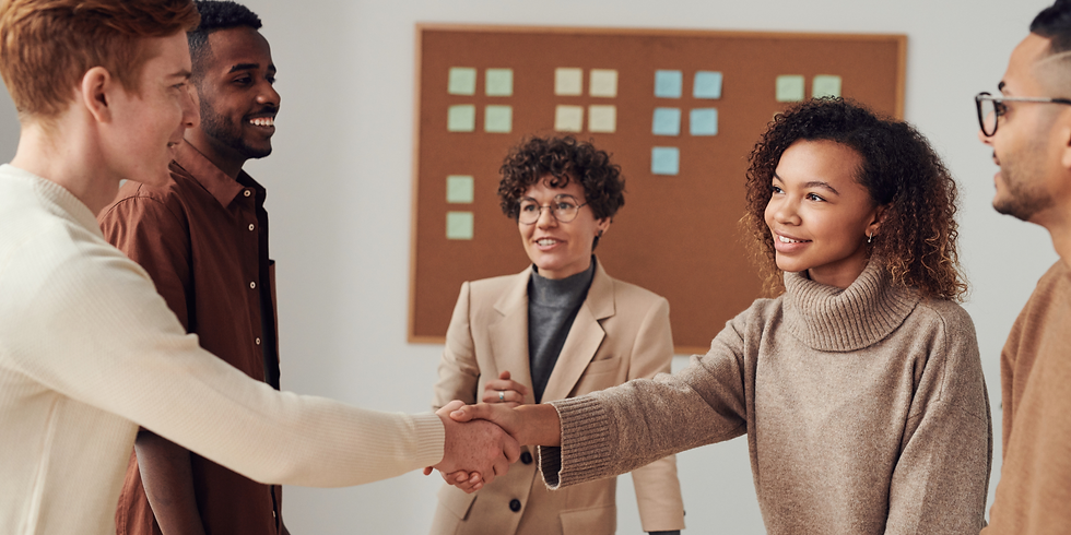 oto de um grupo diversificado de seis profissionais em um escritório. No centro da imagem, um homem branco de cabelo ruivo, vestindo um suéter claro, e uma mulher negra de cabelo cacheado, com um suéter de gola alta bege, apertam as mãos. Os outros quatro colegas os observam sorrindo, criando uma atmosfera positiva e colaborativa. Ao fundo, há um quadro de cortiça com vários post-its.
