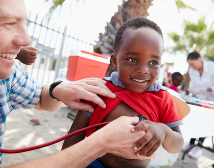 Doctor checks happy boy's heartbeat with stethoscope, wearing red shirt outdoors.