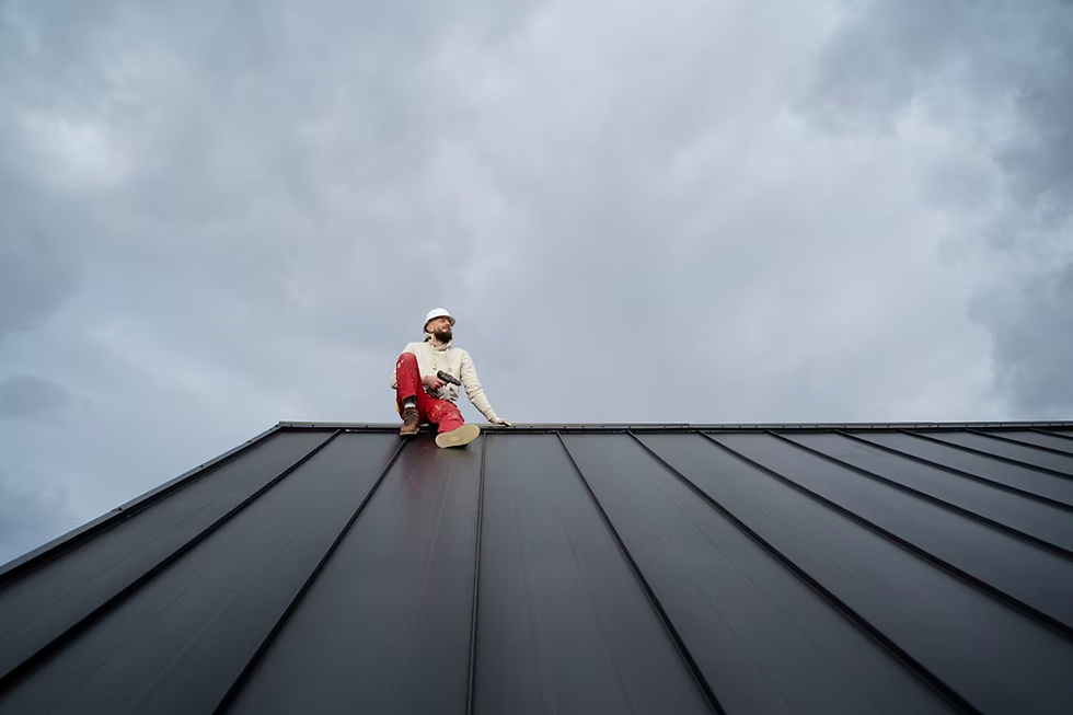 Roof worker sits atop a dark metal roof under a cloudy sky.