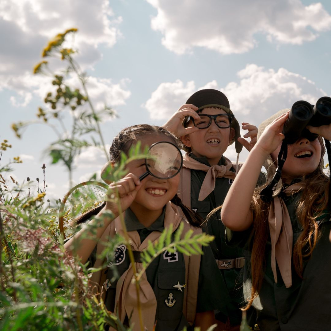 Three children in nature holding binoculars and a magnifying glass.