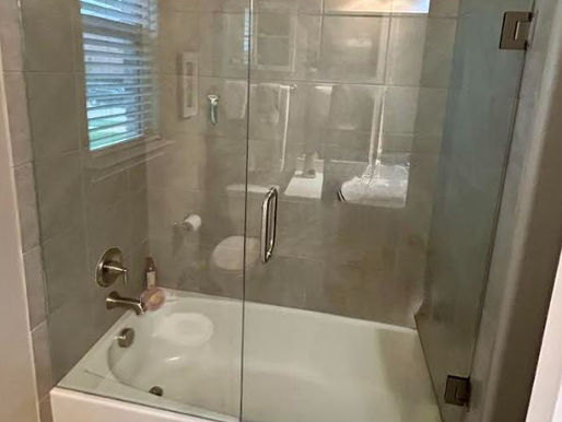 A modern bathroom featuring a clean white bathtub fitted with a custom frameless shower glass enclosure, showcasing a swinging glass door with chrome hardware against gray tiled walls.