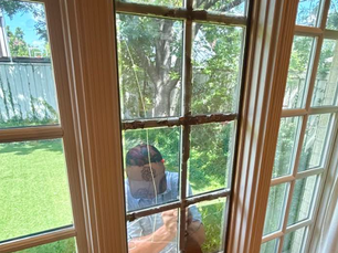 A Collin County Glass window repair technician outside a house seen through a window grid, using a black suction cup tool to remove cracked and broken glass from a wood-framed residential window structure.