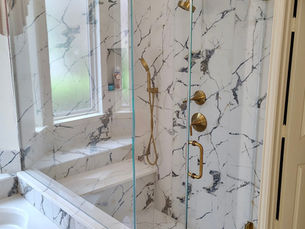 A completed luxury bathroom featuring a frameless three-panel shower enclosure with clear tempered glass and satin brass hardware. The seamless glass showcases the elegant white and gray marble tile work and matching gold fixtures.