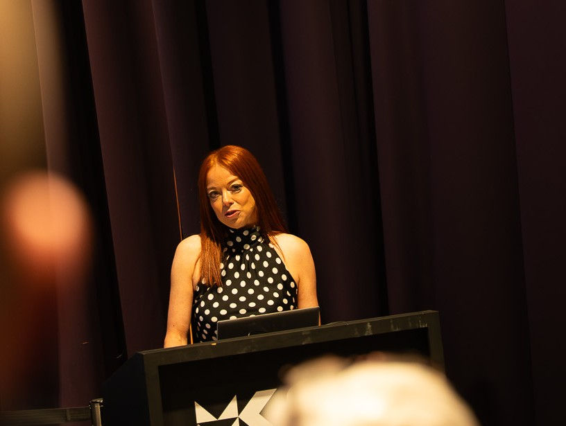 Red-haired woman in polka dot dress speaks at a podium with a "K" logo. Dimly lit stage, dark curtains in the background.