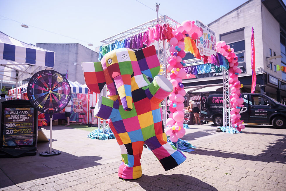 Colorful patchwork elephant mascot at a sunny street festival, with pink balloon arch and Milton Keynes Festival signs in background.