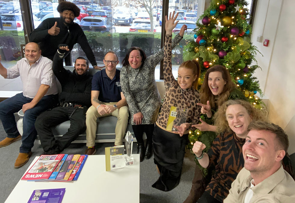 Group of nine people smiling and celebrating indoors by a decorated Christmas tree. Some are seated on a couch, others standing with raised arms.