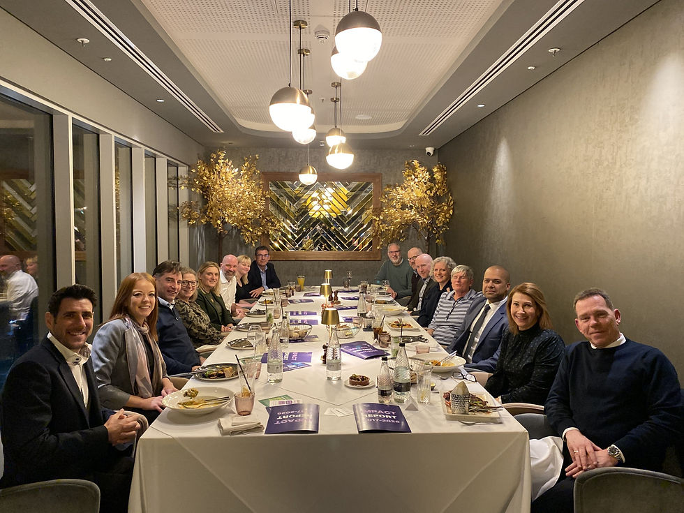 Group of people sitting at a long dinner table enjoying a meal. Elegant setting with gold decor and soft lighting. Papers read "Impact Report."