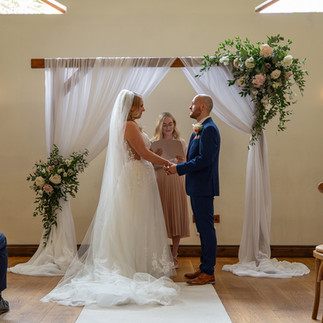 Bride and Groom stood underneath a wood arch with a white cloth and dusky rose flowers and greenery.