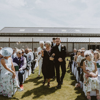 Groomsmen walking with grandmas arm in arm as they throw flower petals down the aisle. Guests sat watching on with cloudy sky.