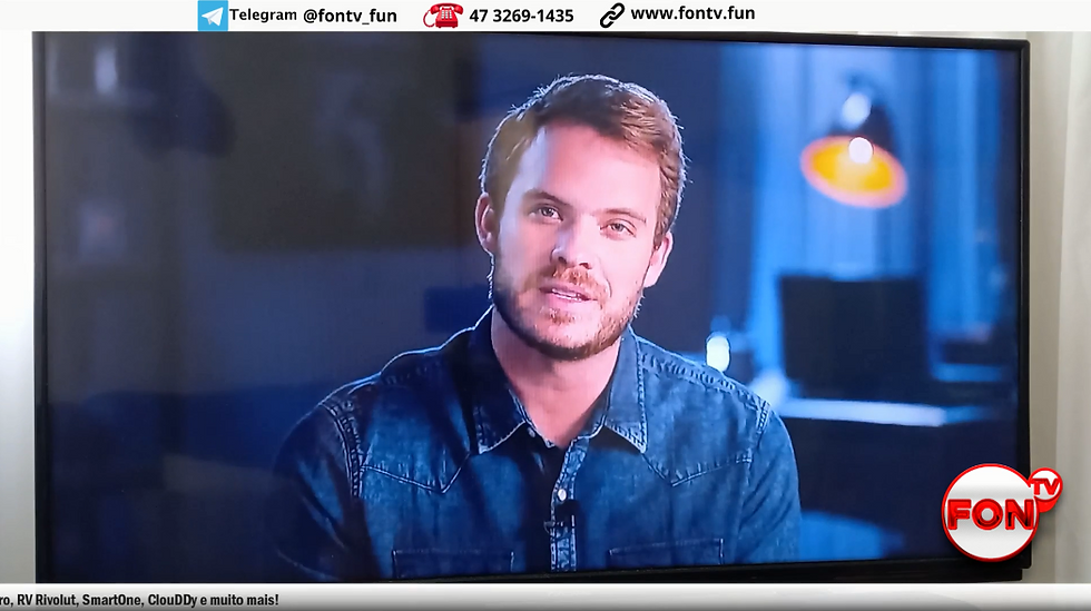 Uma pessoa vestindo uma camisa jeans sentada em frente a uma TV exibindo um logotipo e informações de contato.