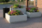Large concrete planters with colorful flowers on brick pavement near a trash can, trees, and a brick building in the background.