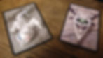 Ghoul card (left) and amulet card (a picture of a skull with antlers, a toothy grin, pointy chin, and one large central green eye; right) on a brown table