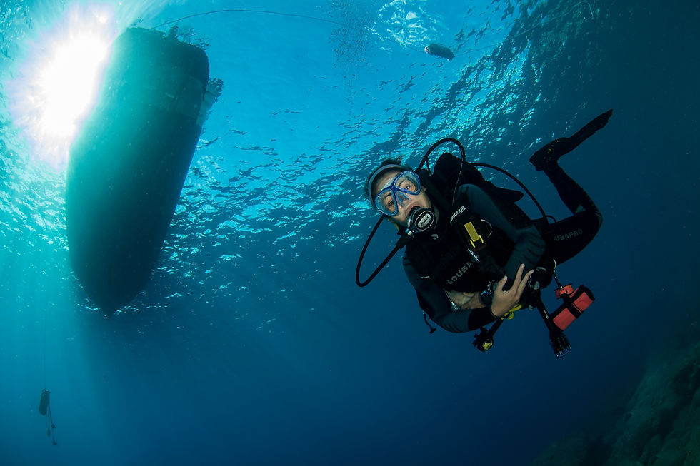 A scuba diver swims underwater near a boat silhouetted against the sun. The ocean is clear blue with rays of light filtering through.