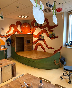 Spielbereich mit einem Schiff aus Holz und einer roten Krake an der Wand, im Vordergrund ein Tisch mit Plastikflasche und Gläsern